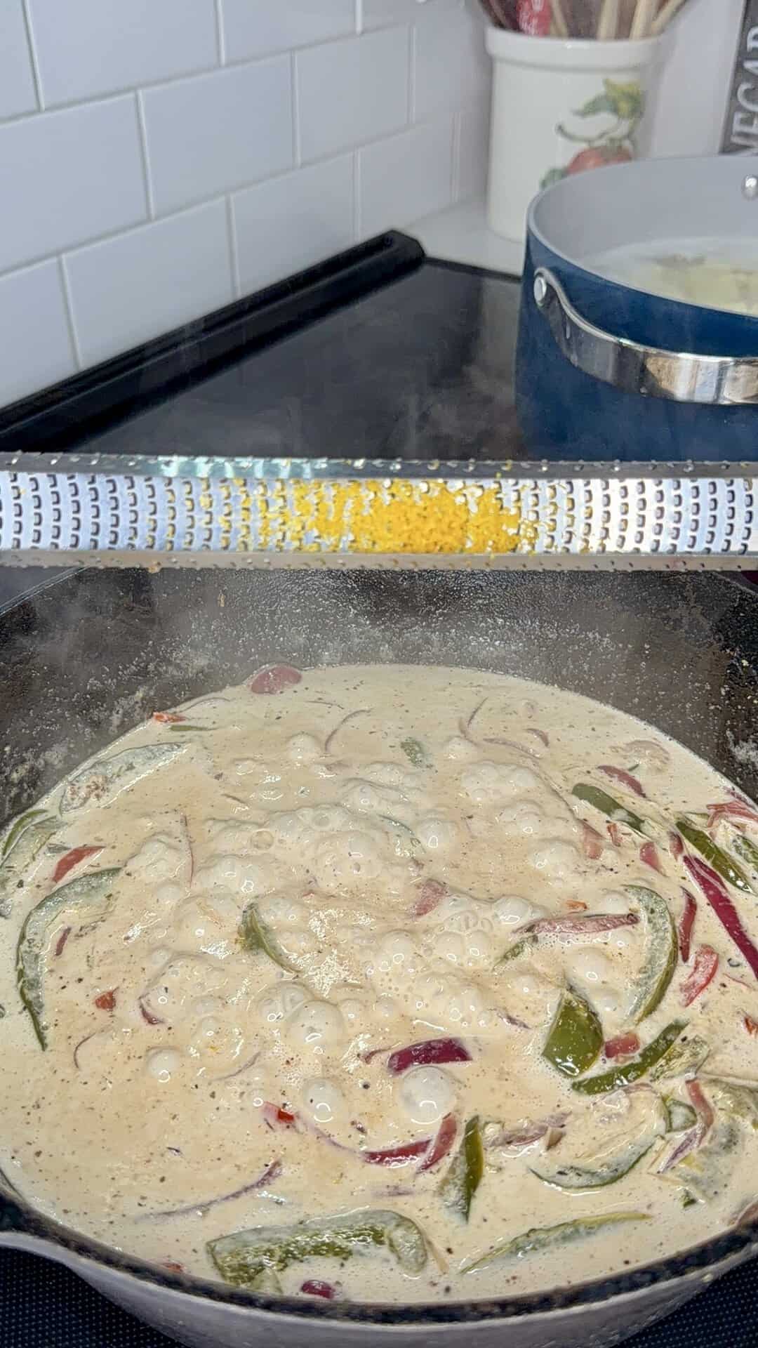 A creamy mixture with sliced peppers cooks in a skillet, while orange zest is being grated over it. A pot sits on the stove in the background.