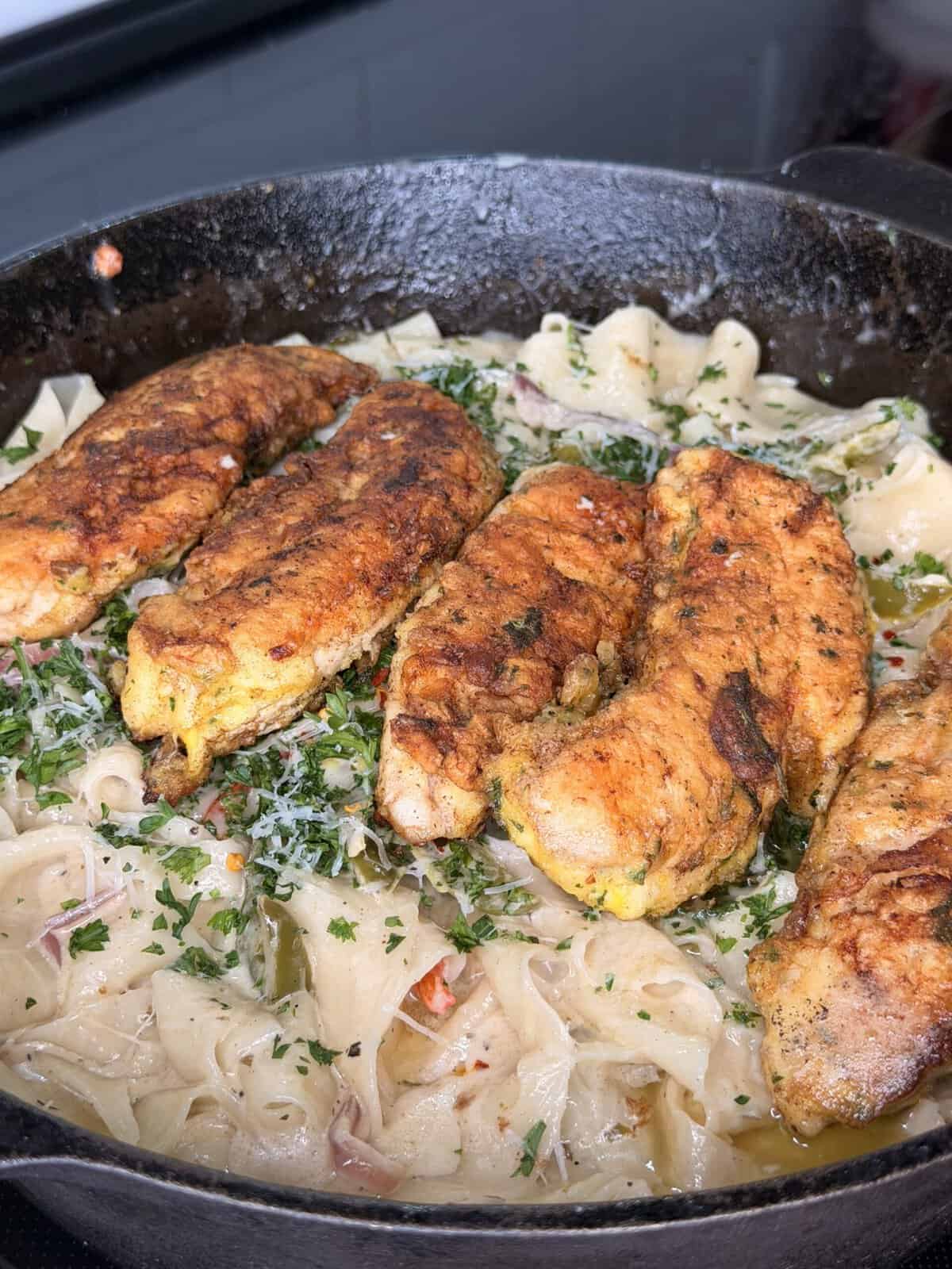 A cast iron skillet filled with creamy fettuccine pasta topped with five seasoned, pan-seared chicken tenders, garnished with chopped herbs and shredded cheese.