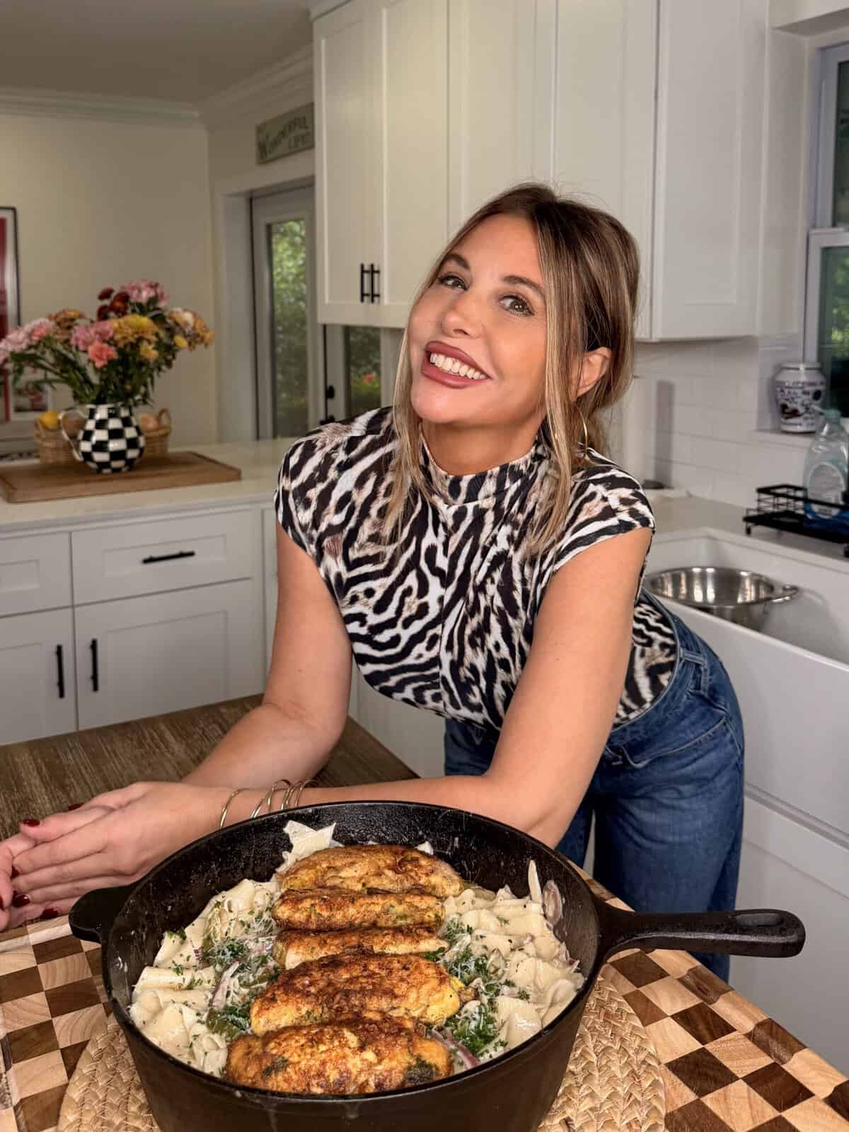 A woman in a zebra-print top smiles in a kitchen, leaning over a table with a skillet of pasta and chicken. Flowers and kitchen decor are in the background.