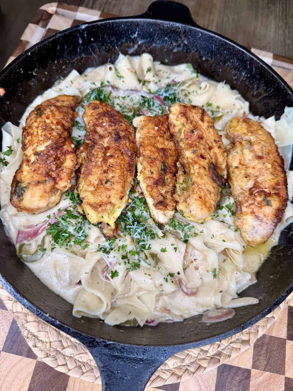 A cast iron skillet filled with creamy pasta topped with five seasoned, golden-brown chicken tenders, garnished with chopped parsley and grated cheese.