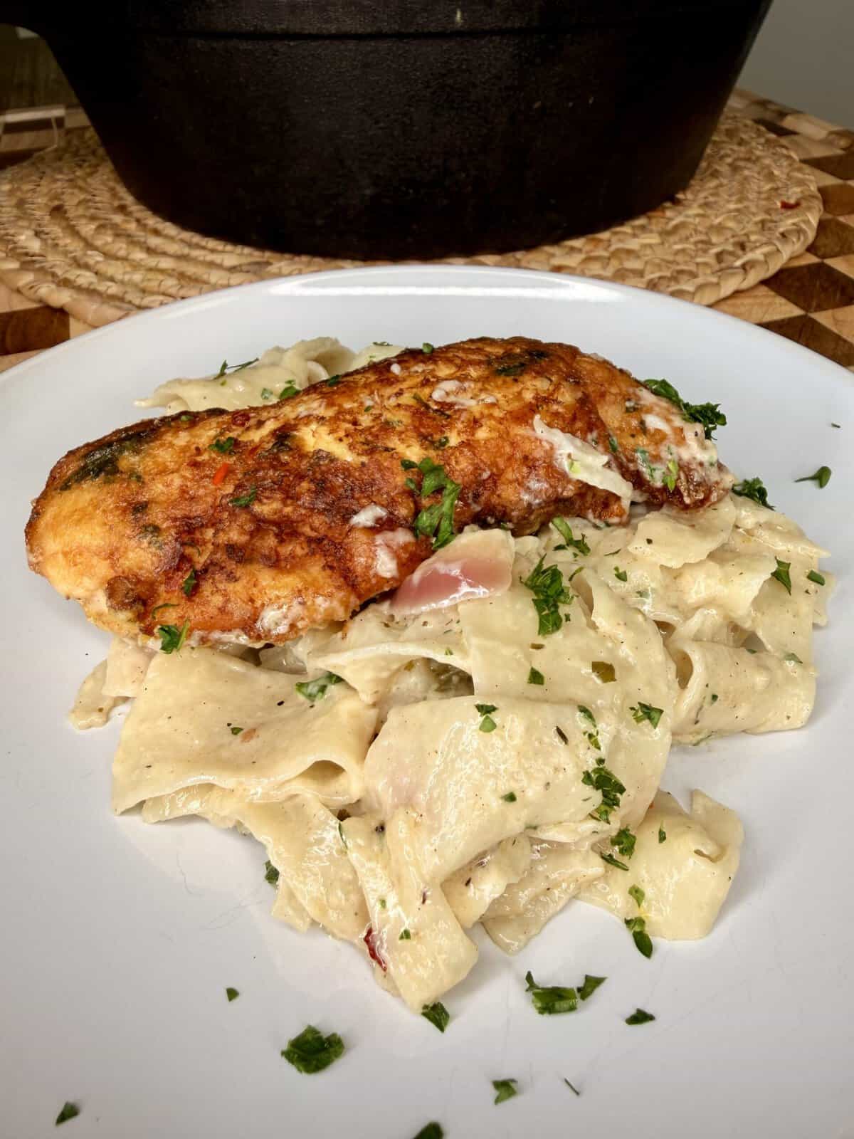 A plate of creamy pasta topped with a golden-brown breaded chicken breast, garnished with chopped parsley. A woven mat and a black pot are visible in the background.