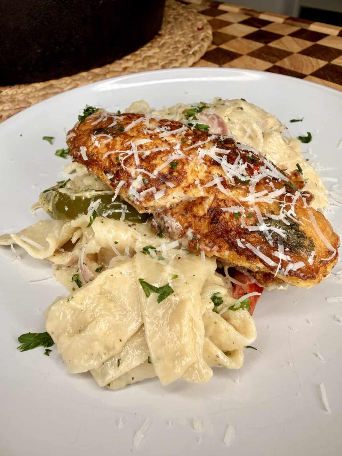 A plate of creamy pasta topped with a breaded, seasoned chicken breast and garnished with shredded cheese and chopped parsley.