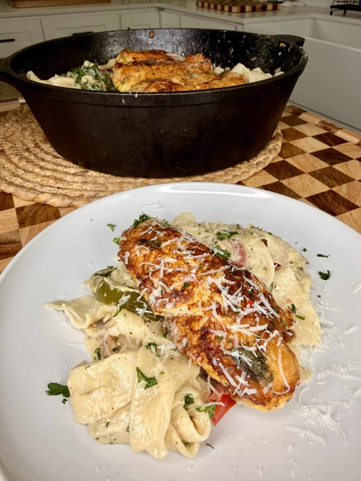 A white plate with creamy pasta, vegetables, and a seasoned, pan-seared fish fillet, topped with grated cheese. In the background, a cast iron skillet with more fish and pasta sits on a woven placemat.