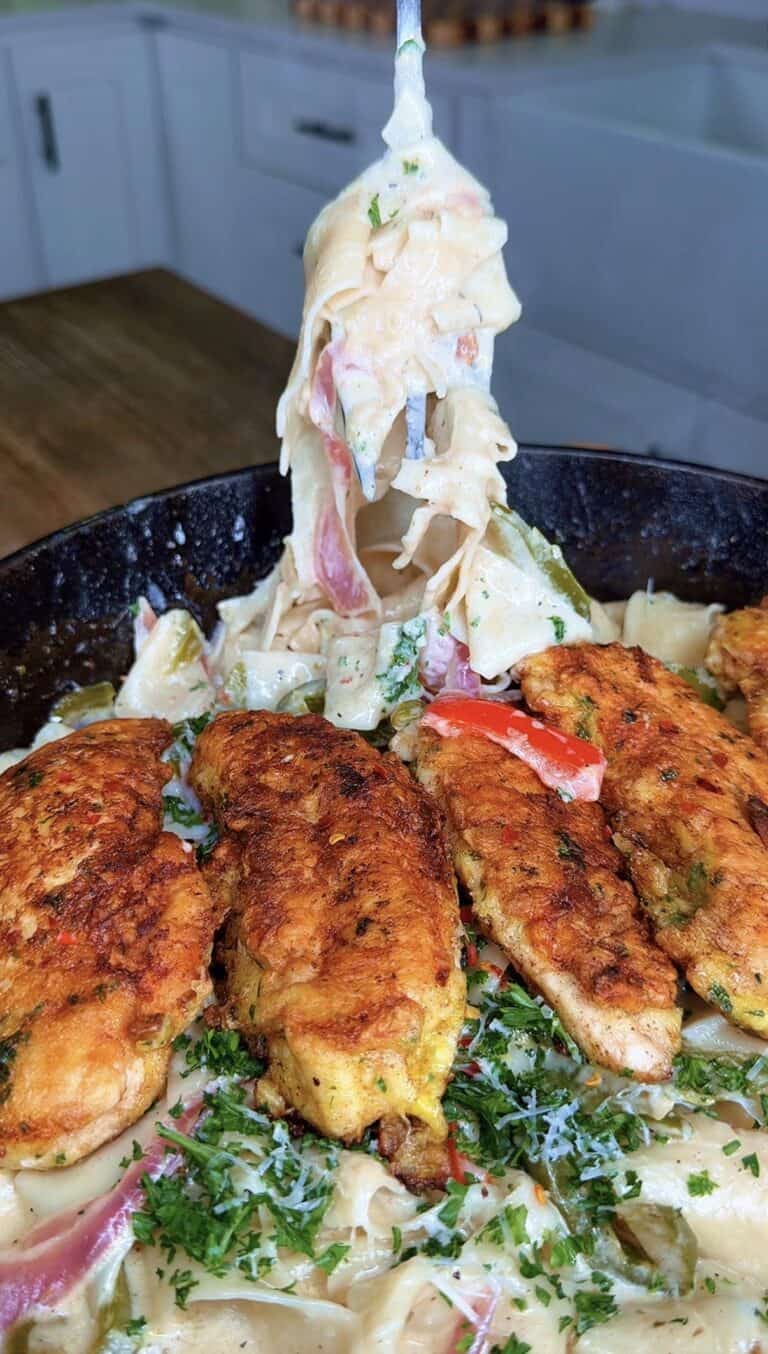 A fork lifts creamy fettuccine pasta above a skillet filled with seasoned, golden-brown chicken strips, colorful bell peppers, and sprinkled herbs, set on a wooden table in a kitchen.