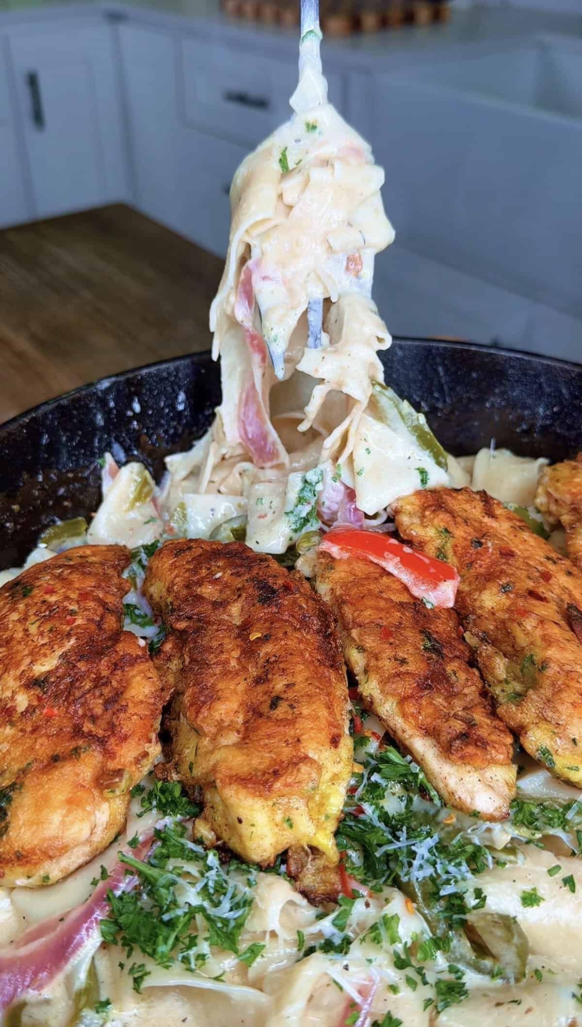 A fork lifts creamy fettuccine pasta above a skillet filled with seasoned, golden-brown chicken strips, colorful bell peppers, and sprinkled herbs, set on a wooden table in a kitchen.