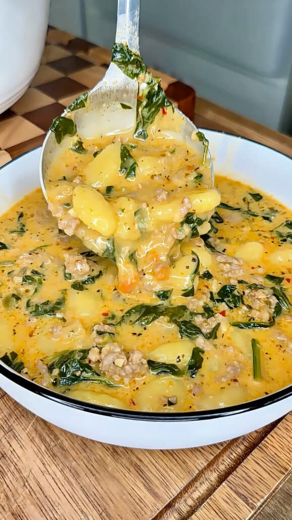A ladle serves creamy soup with gnocchi, sausage, kale, and herbs into a white bowl. The soup is thick, golden, and speckled with seasonings, with visible greens and chunks of sausage.