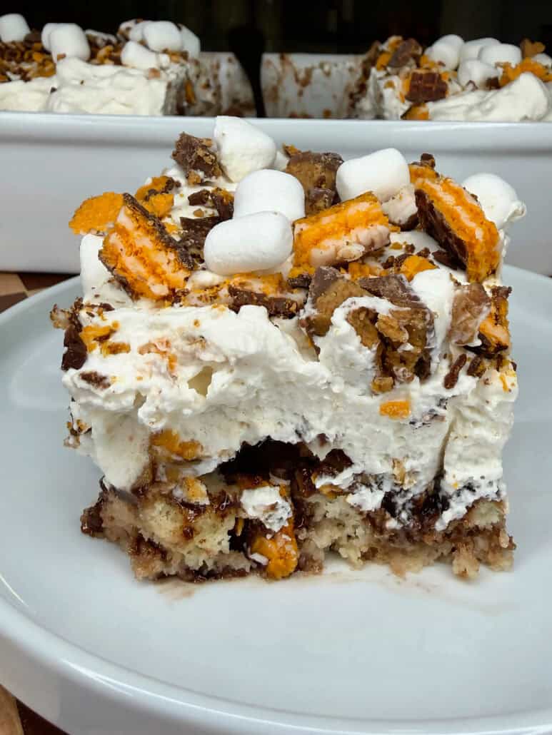 A close-up of a dessert bar topped with whipped cream, marshmallows, chocolate, and orange candy pieces, served on a white plate. A baking dish with more dessert is in the background.