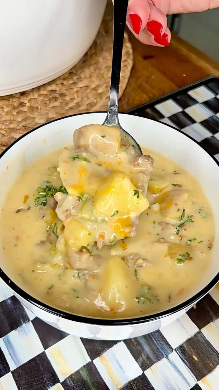 A hand holding a spoonful of creamy soup with potatoes, vegetables, and herbs over a bowl, all set on a checkered placemat.