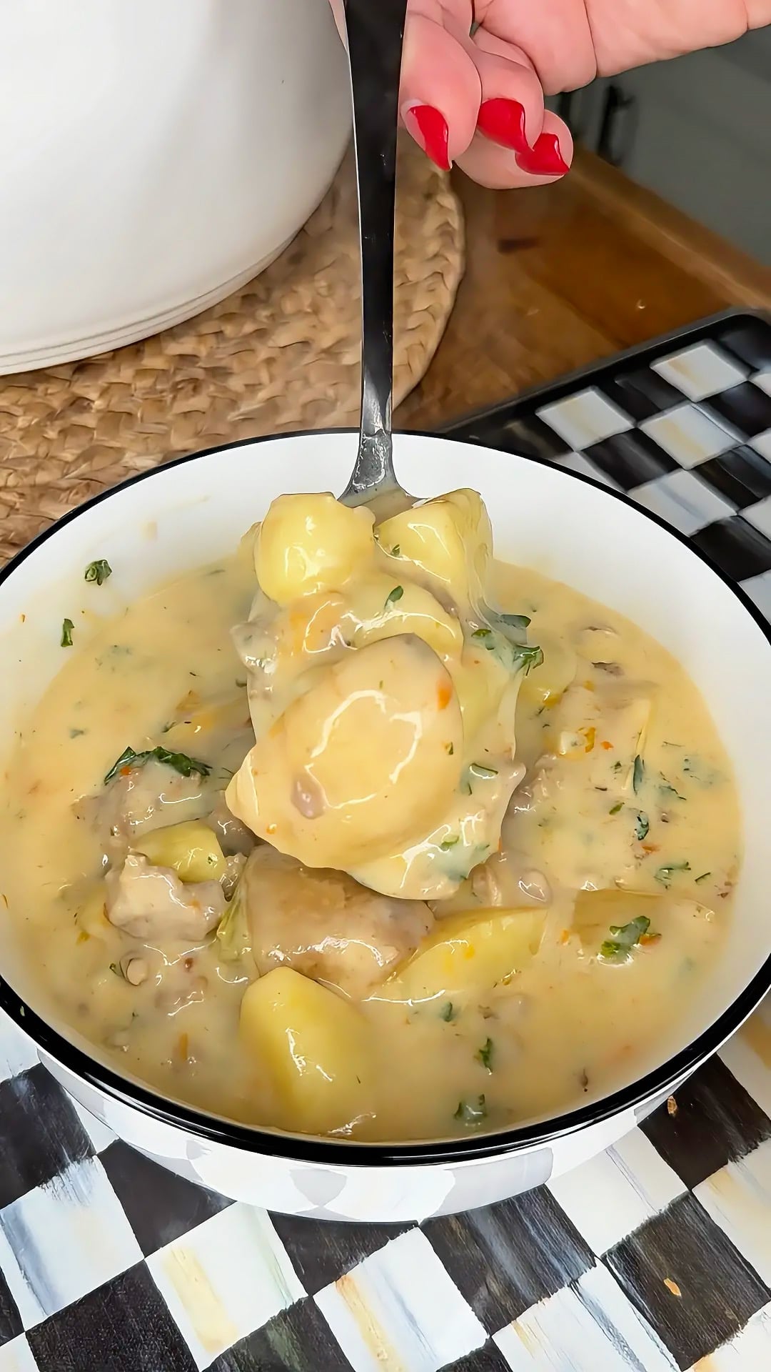 A hand holds a spoonful of creamy potato soup with herbs and chunks of potatoes over a bowl, set on a checkered placemat.
