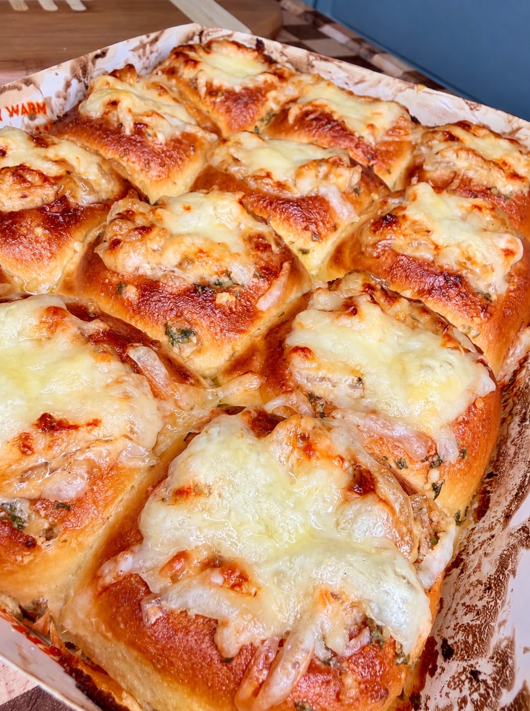 A close-up of golden, baked bread rolls topped with melted cheese and herbs, arranged closely together in a tray.