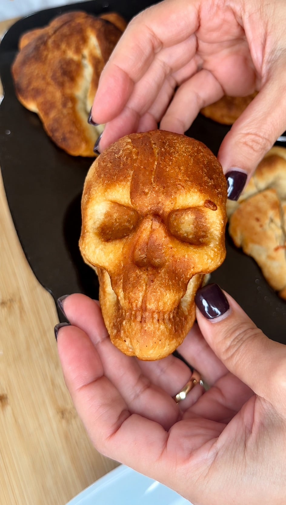 Hands holding a baked bread roll shaped like a human skull.