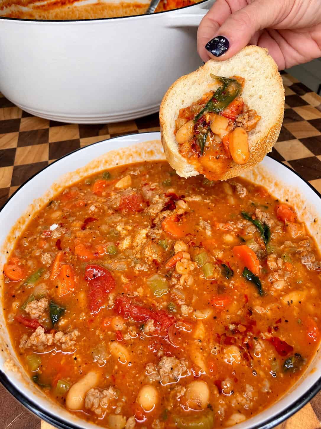 A hand holds a slice of bread dipped into a bowl of tomato-based soup with sausage, beans, and vegetables.