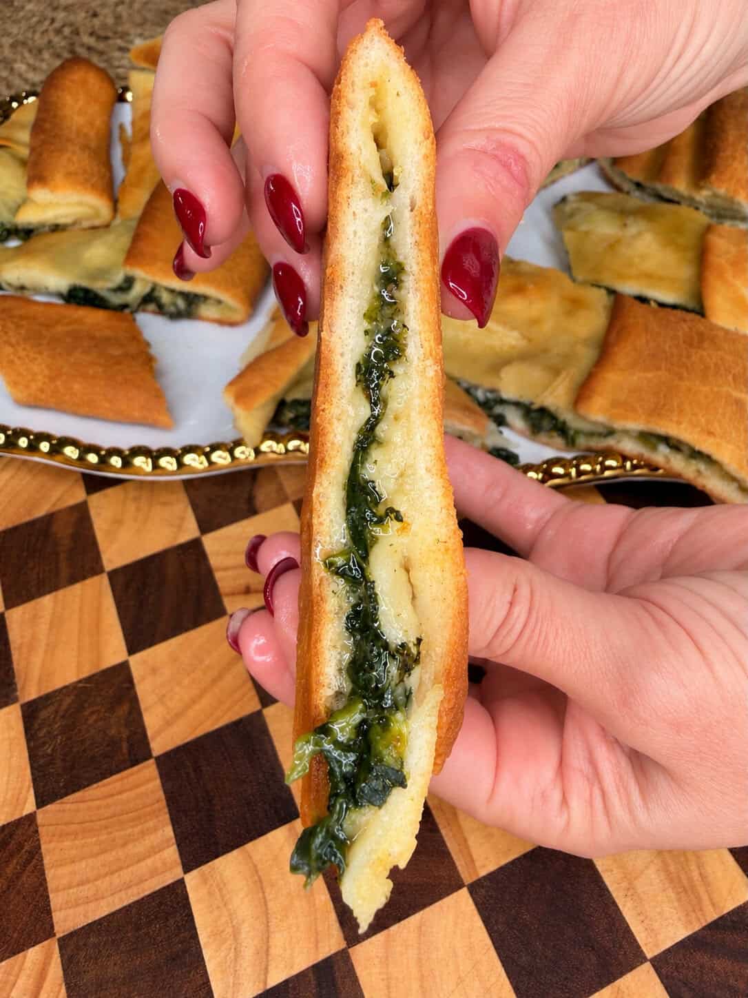 A hand holds a piece of flatbread filled with melted cheese and spinach, with more pieces on a platter in the background.