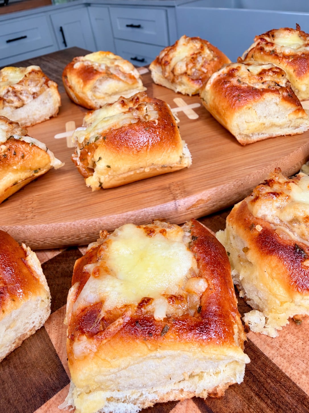 Golden, cheesy bread rolls with melted cheese on top are arranged on a wooden board in a kitchen setting. The rolls appear soft, fluffy, and slightly browned around the edges.