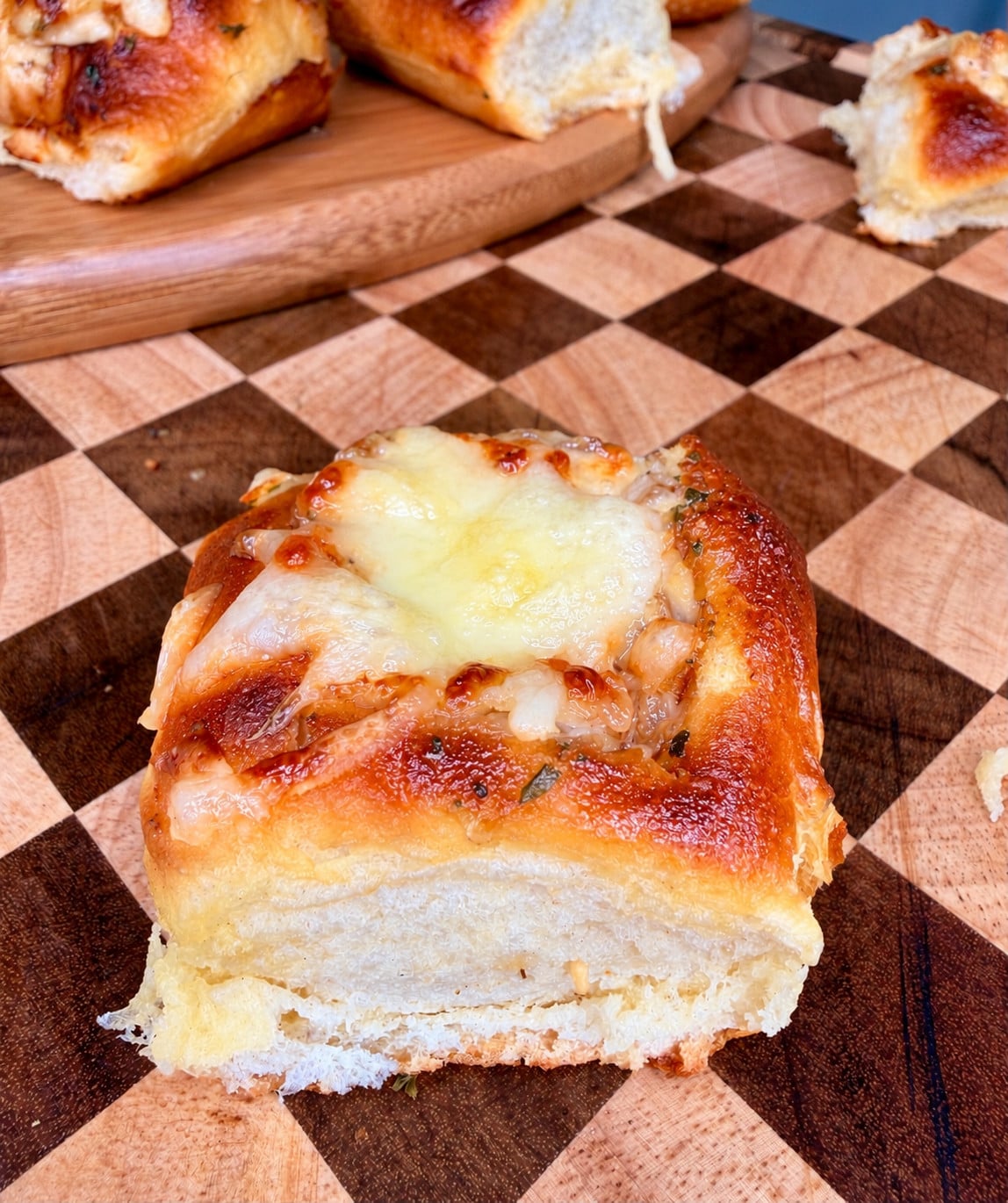 A close-up of a soft, golden-brown bread roll topped with melted cheese, resting on a wooden checkered cutting board. Other rolls are visible in the background.