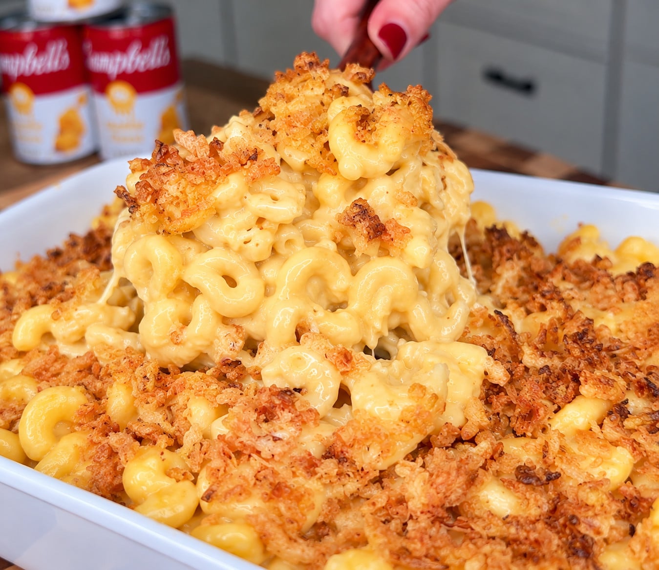 A serving spoon lifts a portion of creamy macaroni and cheese topped with toasted breadcrumbs from a baking dish.
