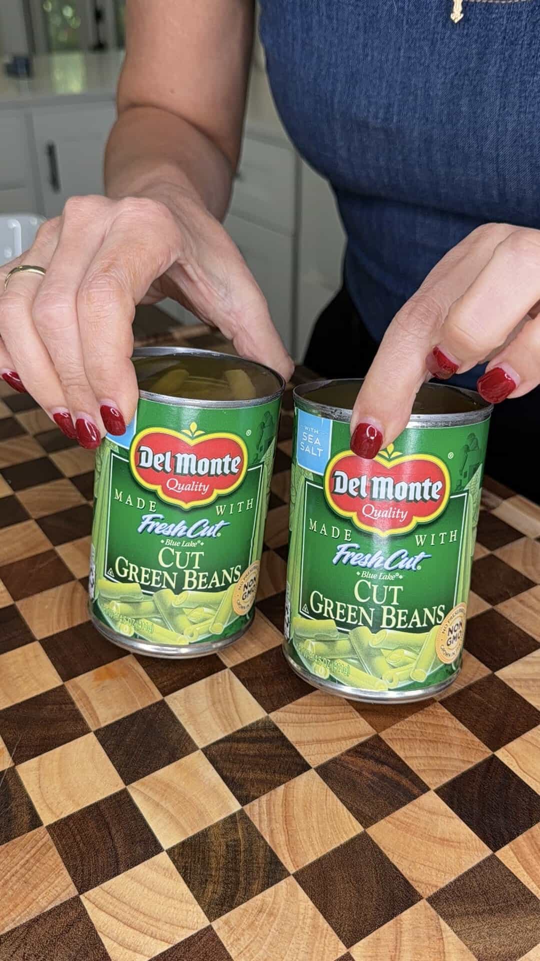 A person holds two cans of Del Monte Cut Green Beans on a checkerboard wooden surface.