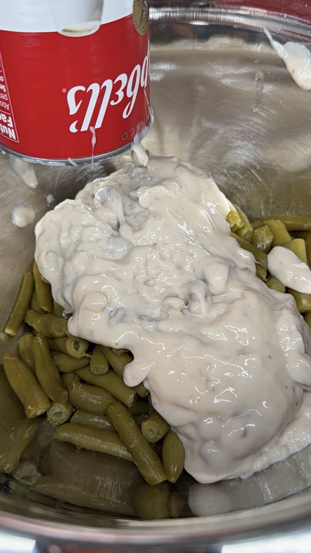 Cream of mushroom soup being poured from a red Campbells can into a bowl of green beans.