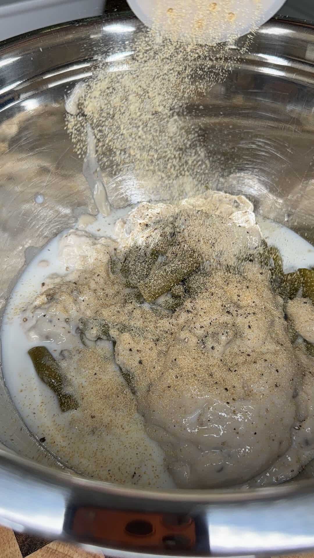 A spice or seasoning powder being sprinkled into a stainless steel mixing bowl containing a creamy mixture and green pepper pieces.