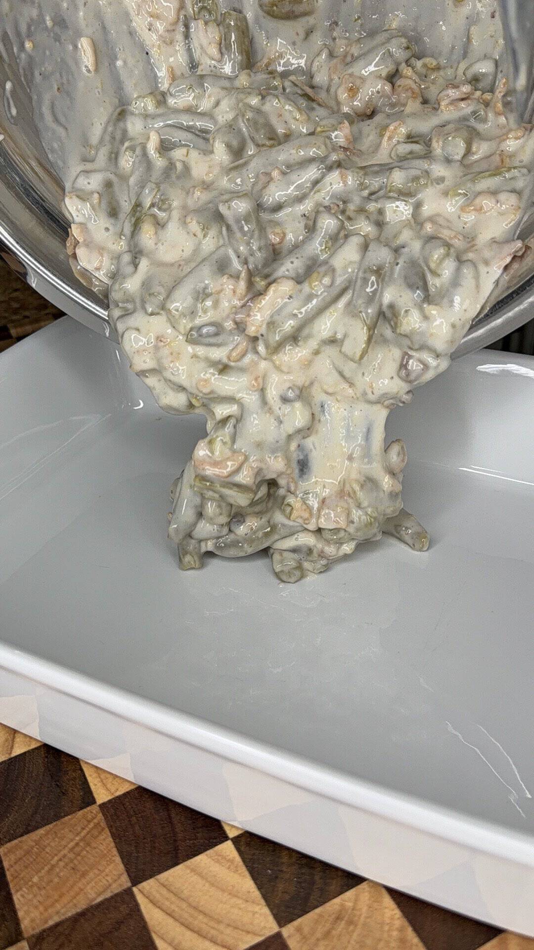 Creamy green bean casserole being poured from a metal bowl into a white ceramic baking dish.