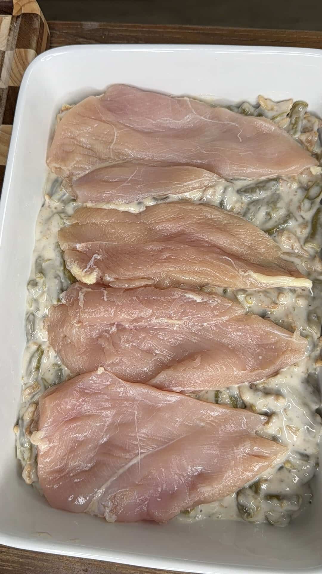 Four raw chicken breasts arranged on top of green beans in a creamy sauce in a baking dish.
