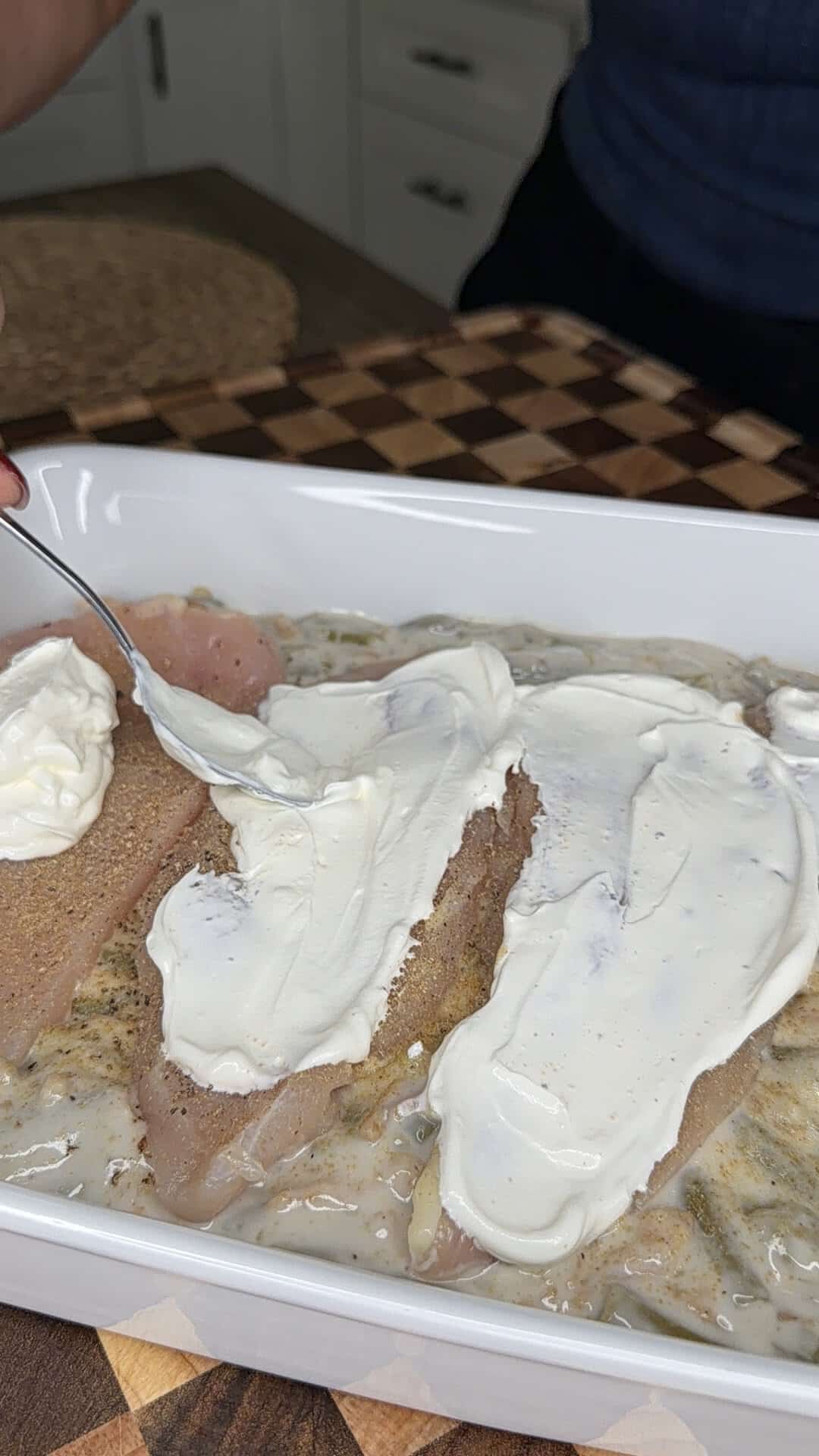 A person uses a spoon to spread a layer of creamy white topping onto raw chicken breasts in a baking dish.