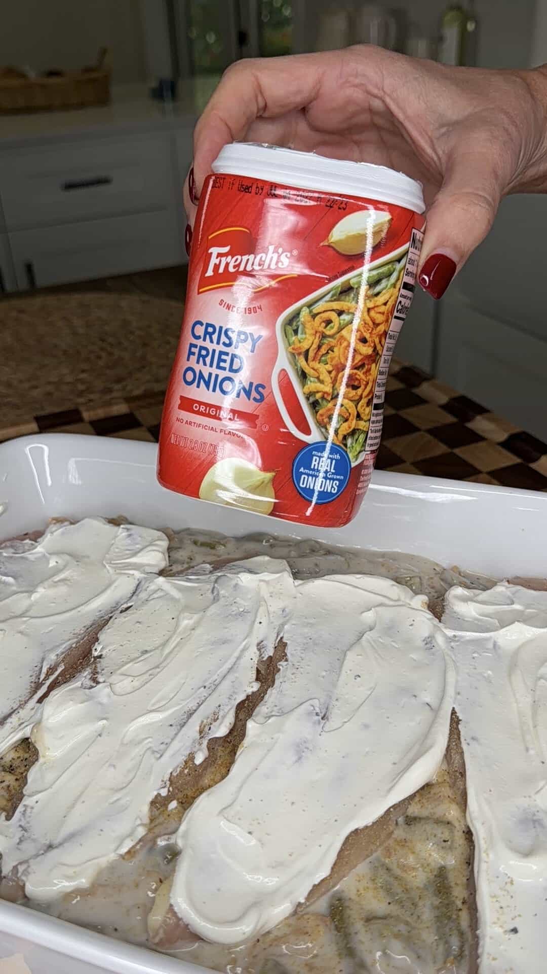A hand holds a container of Frenchs Crispy Fried Onions over a baking dish containing meat covered in a white sauce.
