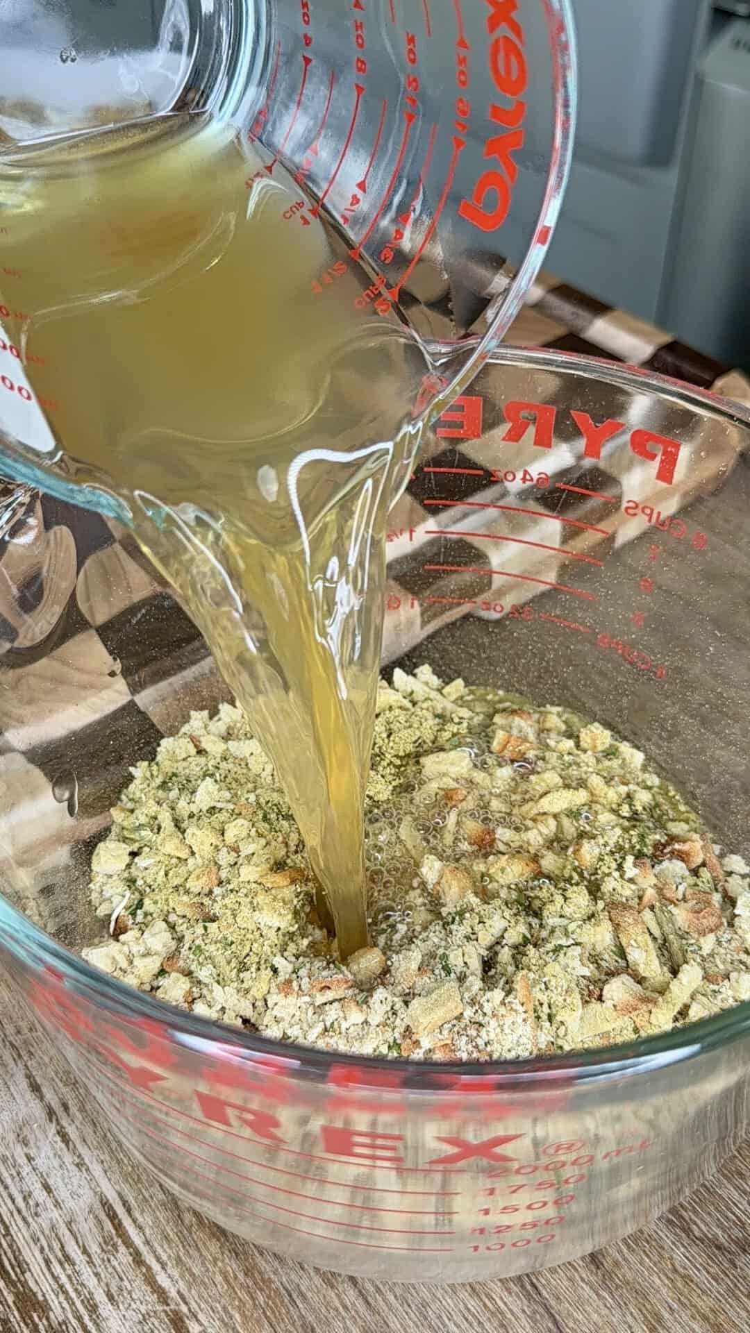Broth being poured from a glass measuring cup into a larger glass bowl filled with herb-seasoned breadcrumbs.