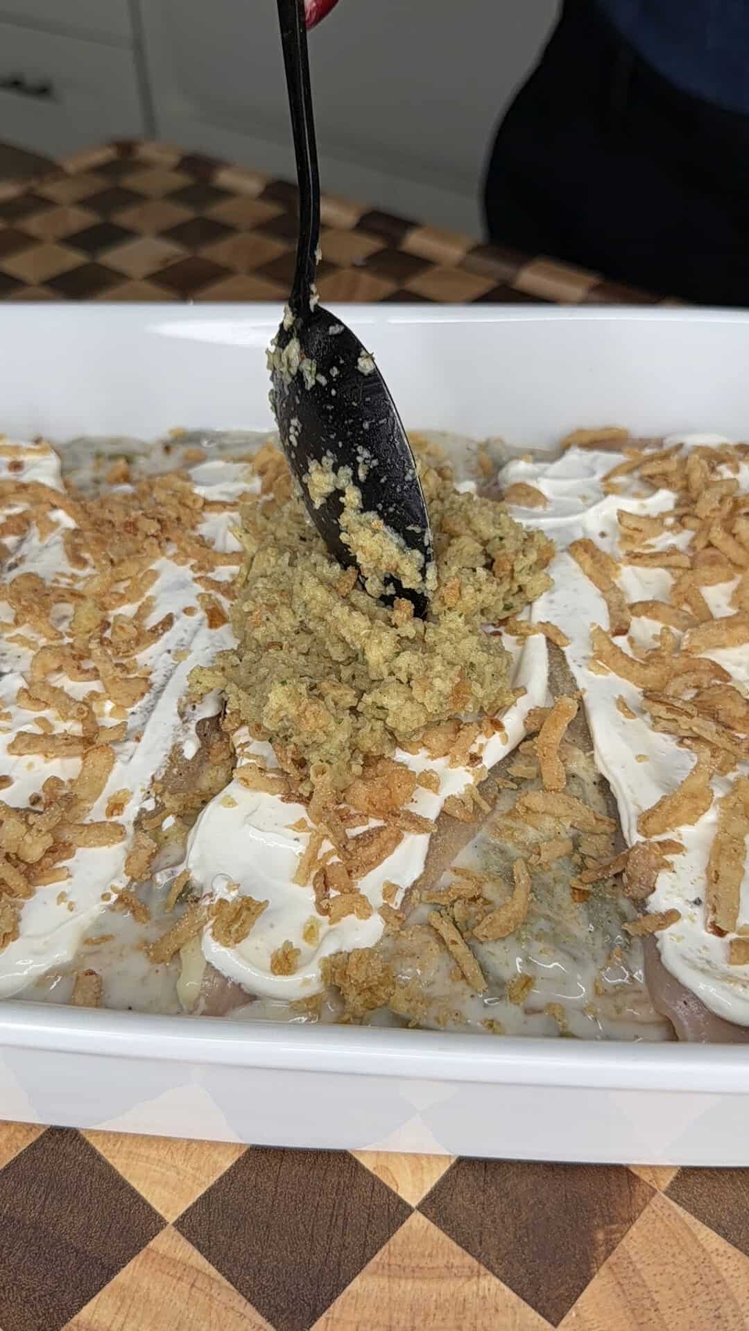 A black spoon adds stuffing to a baking dish containing chicken, creamy sauce, and crispy fried onions.