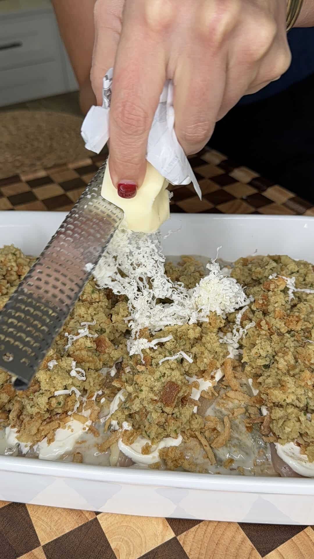 A hand uses a handheld grater to shred a block of cold butter over a casserole topped with breadcrumb stuffing.