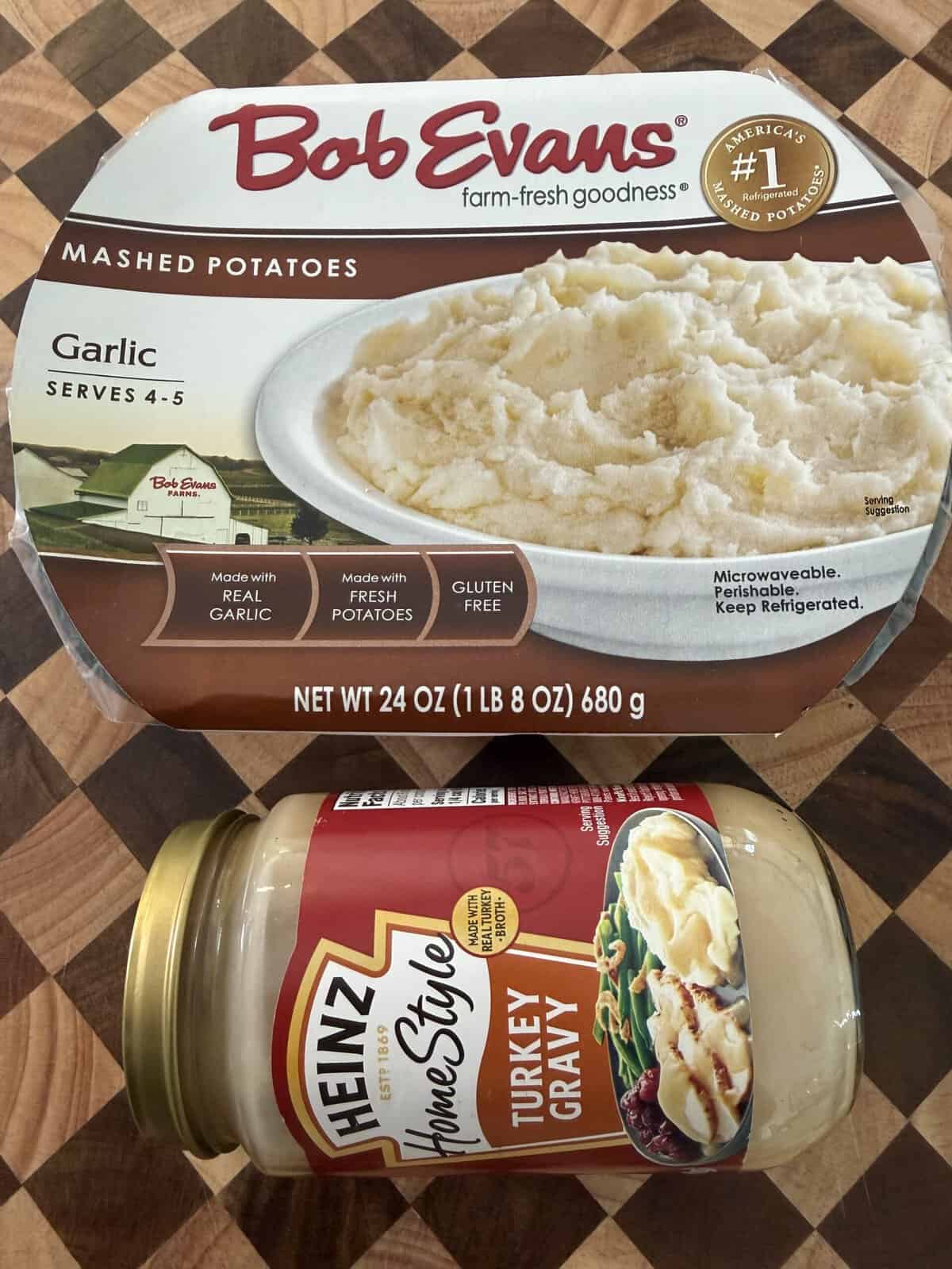 A container of Bob Evans garlic mashed potatoes sitting above a jar of Heinz HomeStyle turkey gravy on a wooden surface.