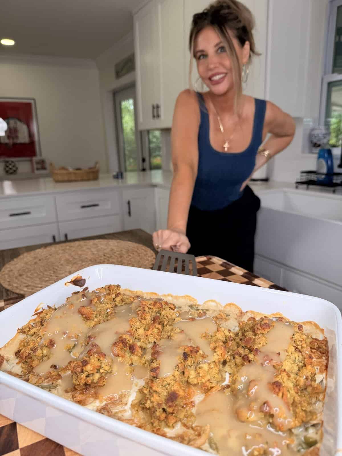 A woman in a blue tank top stands behind a white baking dish filled with a chicken and stuffing casserole topped with gravy.