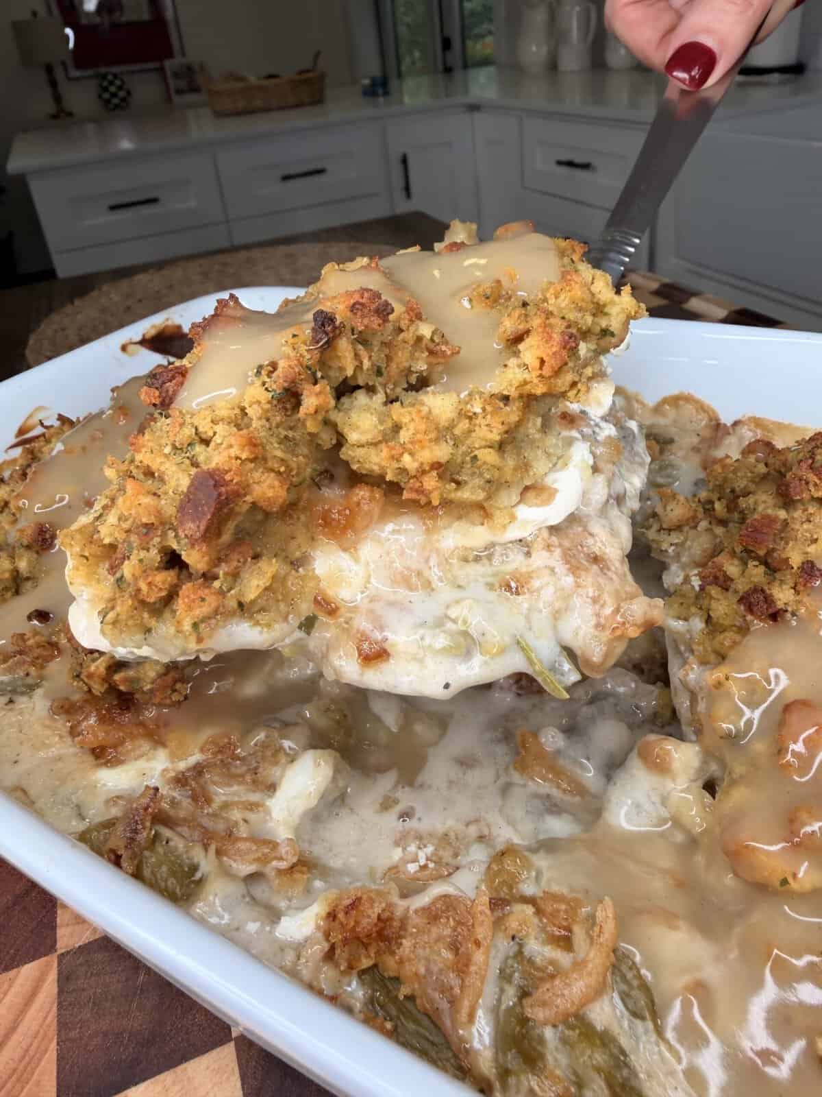 A serving spoon lifts a portion of creamy casserole topped with herb stuffing and brown gravy from a baking dish.