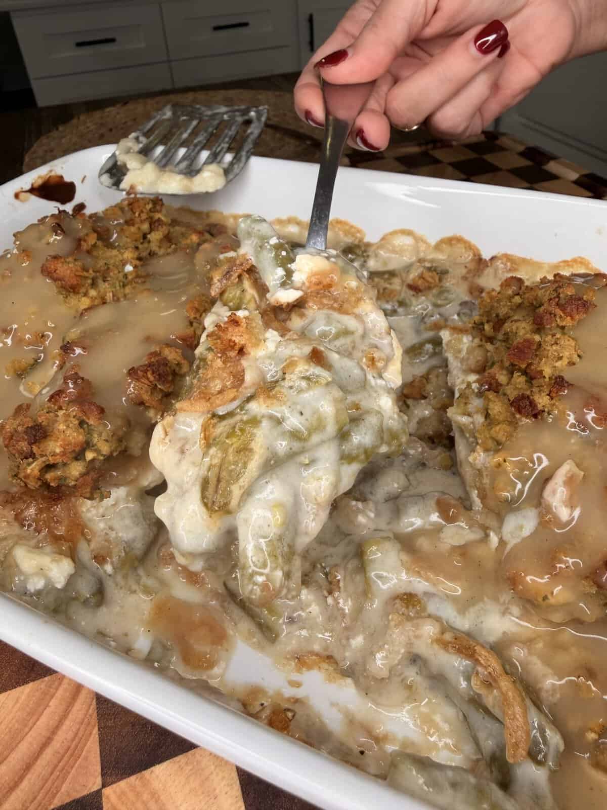 A hand holds a serving spoon lifting a portion of green bean casserole topped with stuffing and gravy from a baking dish.