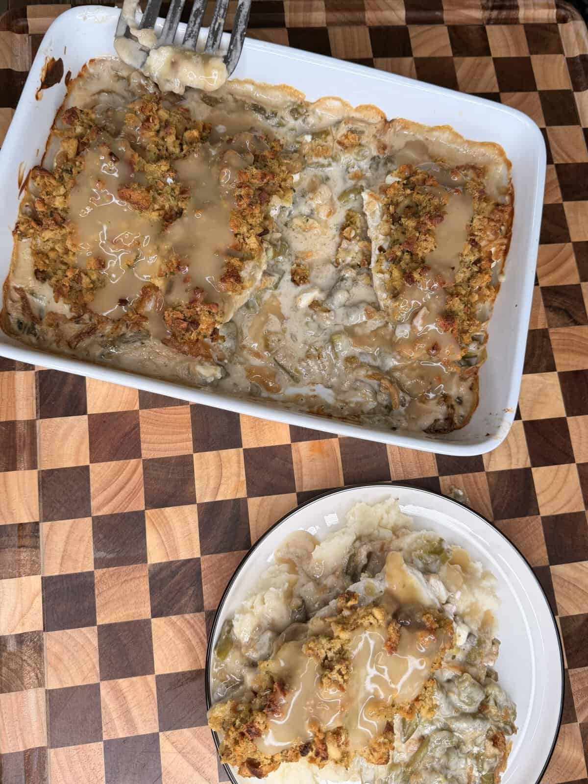 A white casserole dish of Thanksgiving Chicken & Stuffing Supreme, topped with gravy, next to a plate with a serving of the same dish.