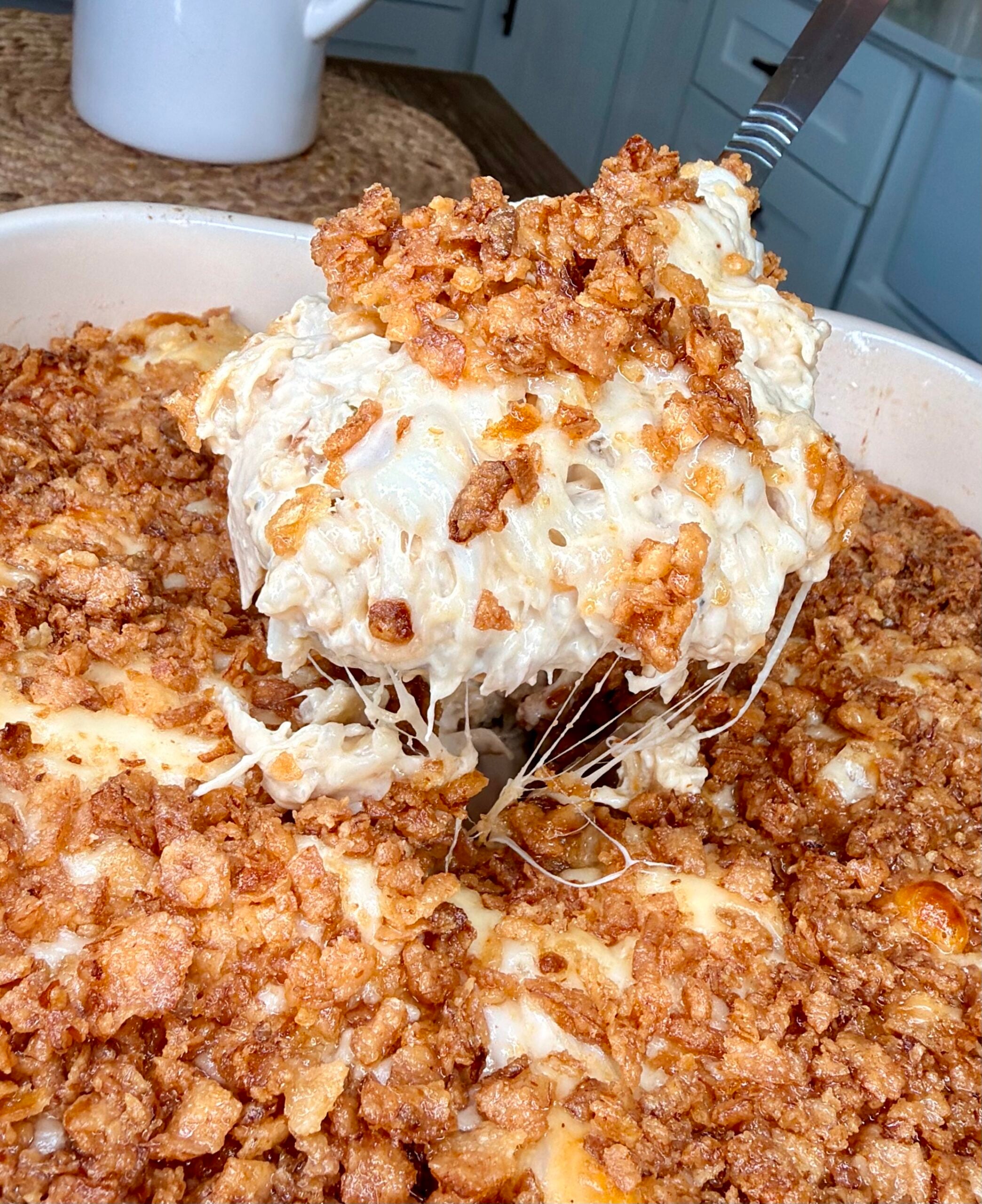 A serving spoon lifts a portion of cheesy, creamy casserole topped with crispy fried onions from a baking dish.