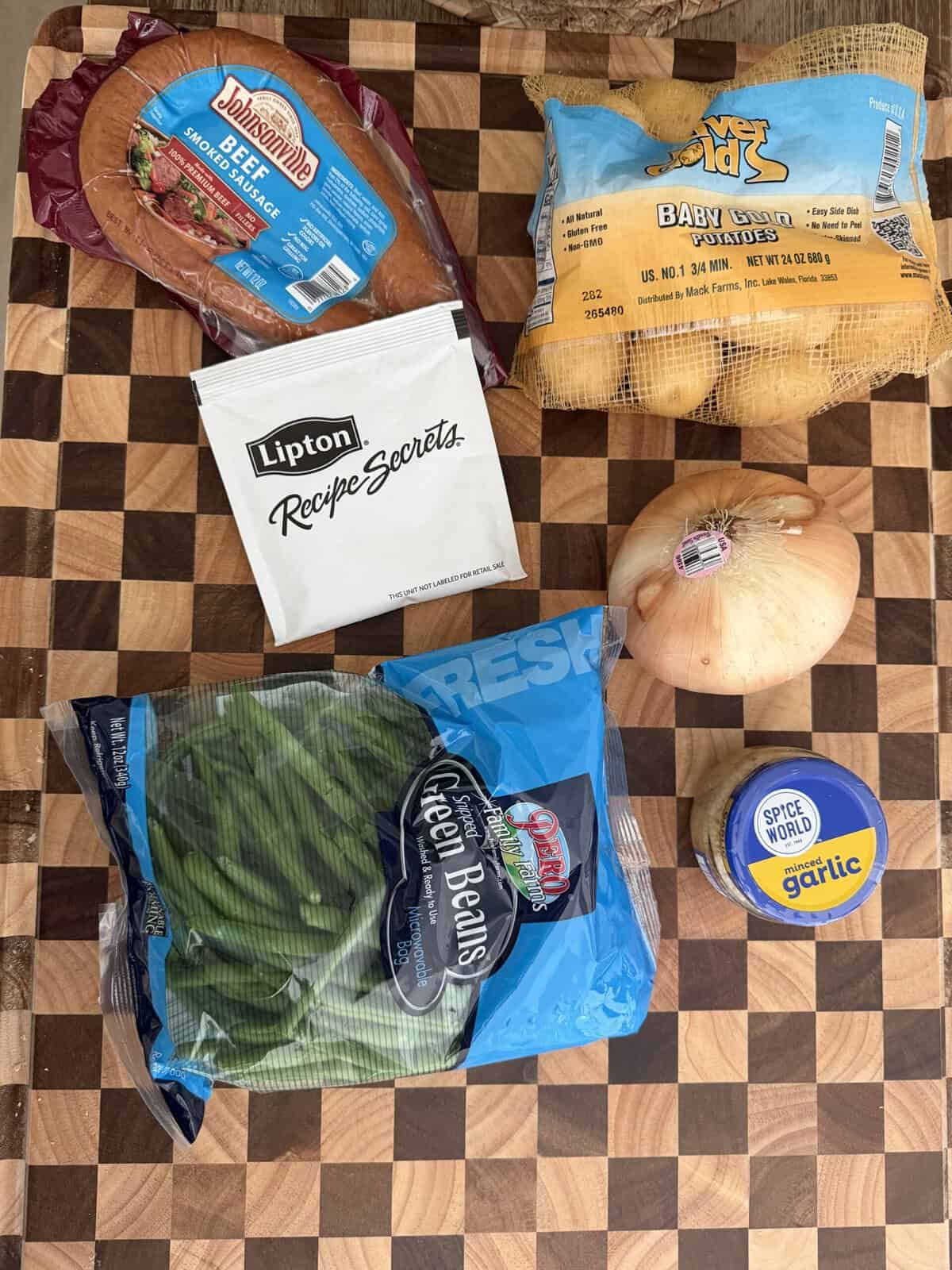 Ingredients for a meal arranged on a cutting board, including a package of Johnsonville beef smoked sausage, a bag of baby gold potatoes, a packet of Lipton Recipe Secrets, an onion, a bag of green beans, and a jar of minced garlic.