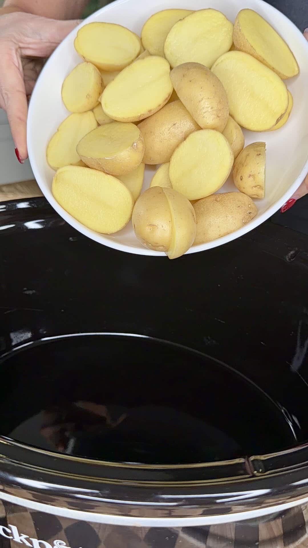 Hands holding a white bowl filled with halved raw potatoes over a slow cooker.