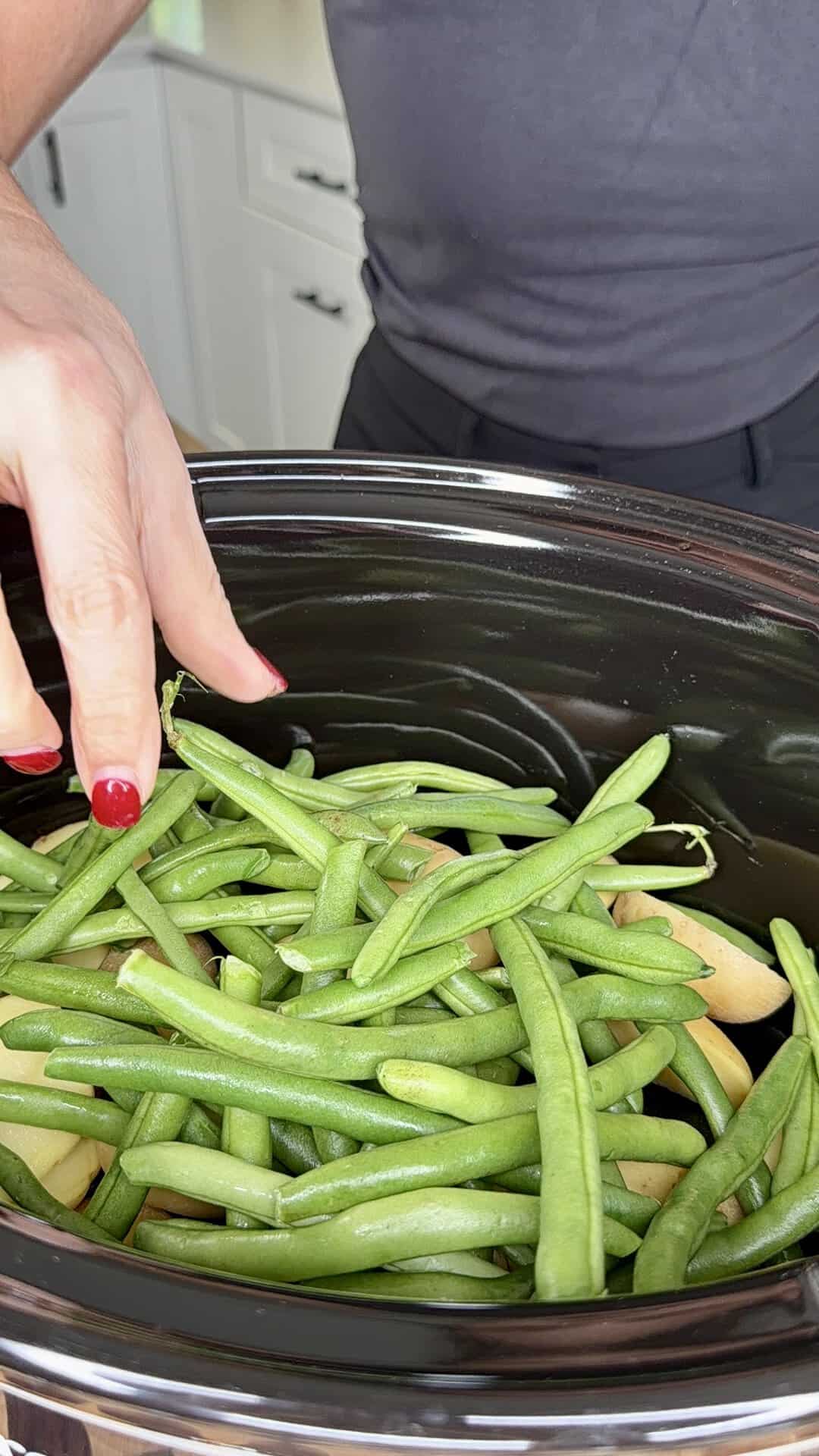 A hand arranging raw green beans and potato slices in a slow cooker.