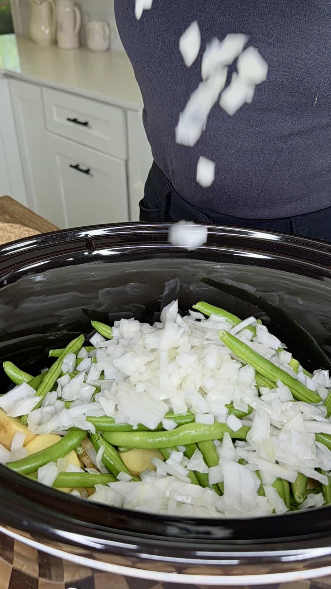 Chopped onions falling into a slow cooker filled with green beans and potato slices.