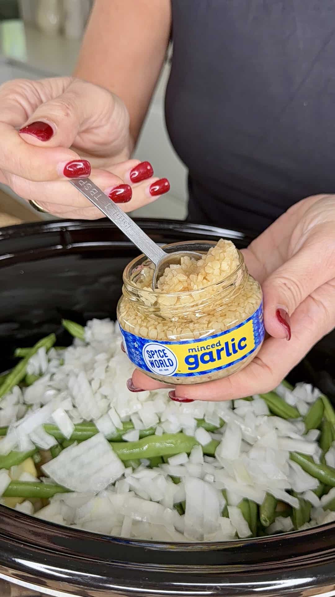 Hands holding a small jar of Spice World minced garlic and a measuring spoon over a slow cooker filled with chopped onions and green beans.