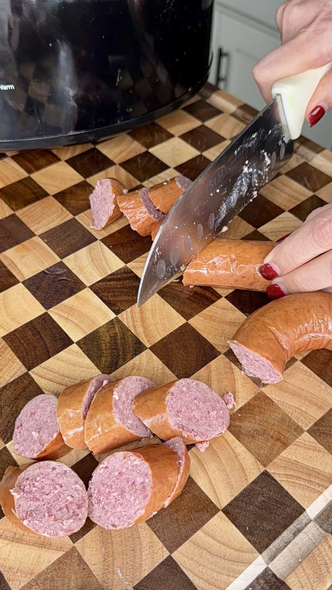 A person using a kitchen knife to slice a ring of sausage on a checkered wooden cutting board.