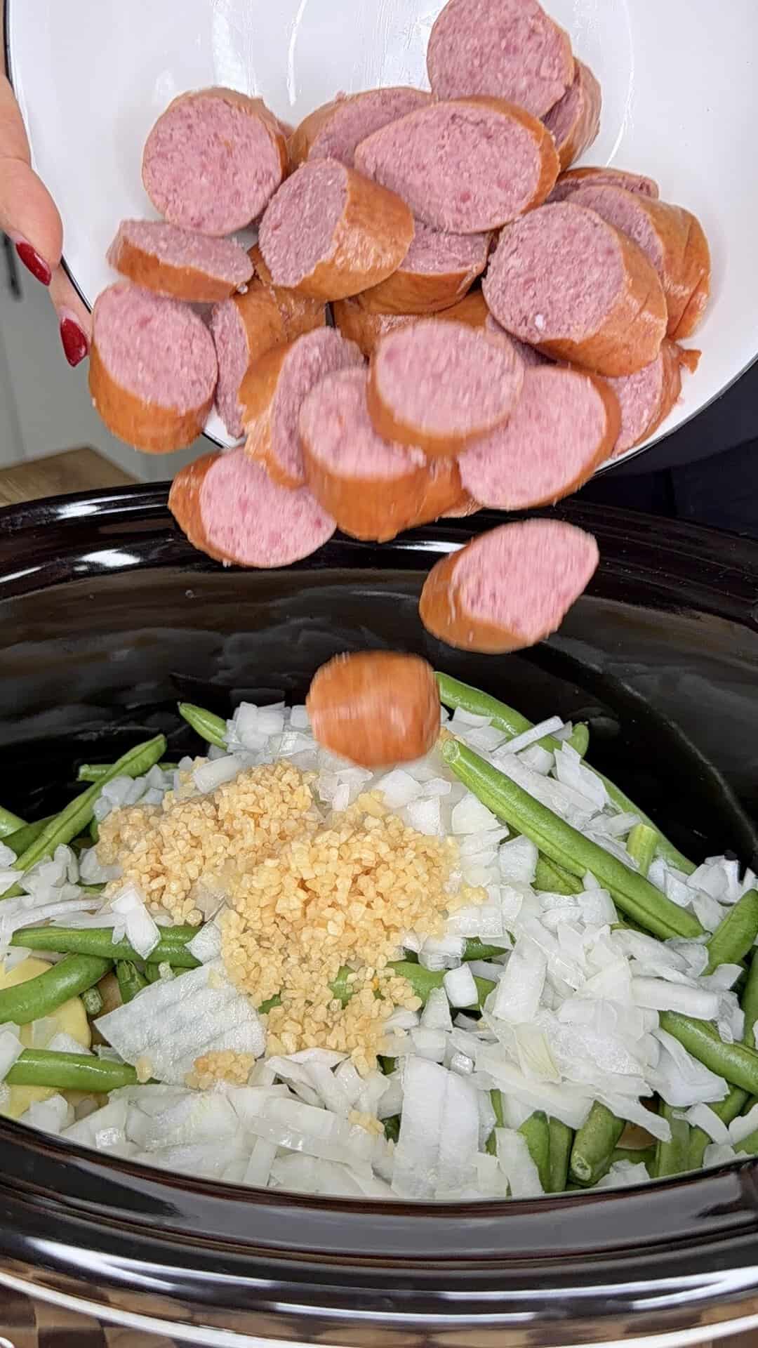 Sliced sausage being poured from a plate into a slow cooker containing green beans, chopped onions, and minced garlic.