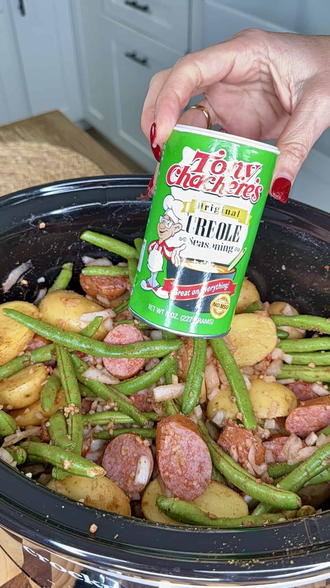 A hand holds a container of Tony Chacheres Original Creole Seasoning over a slow cooker filled with green beans, sliced sausage, and potatoes.