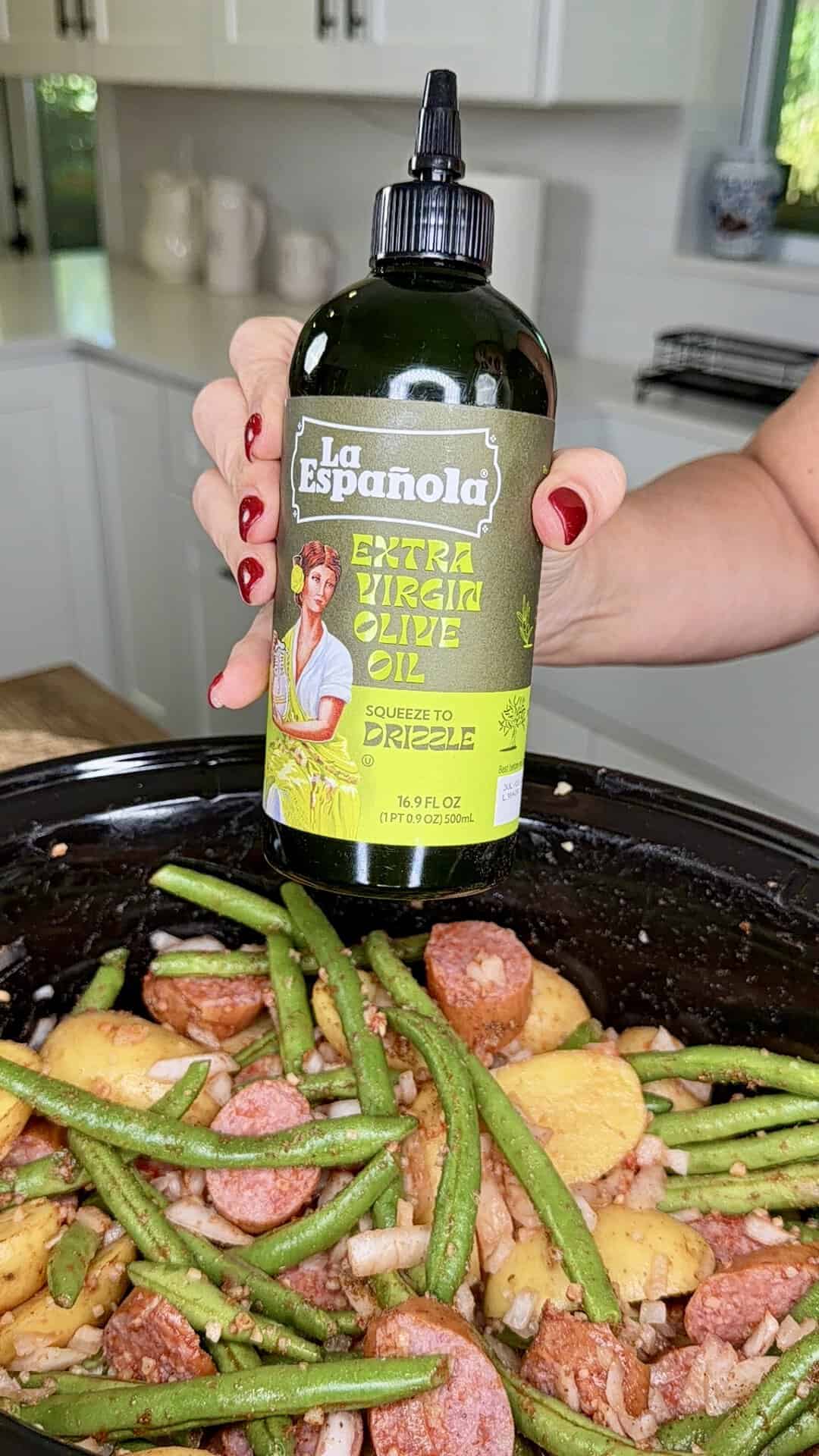 A hand holds a squeeze bottle of La Española extra virgin olive oil over a slow cooker filled with green beans, sliced sausage, and potatoes.