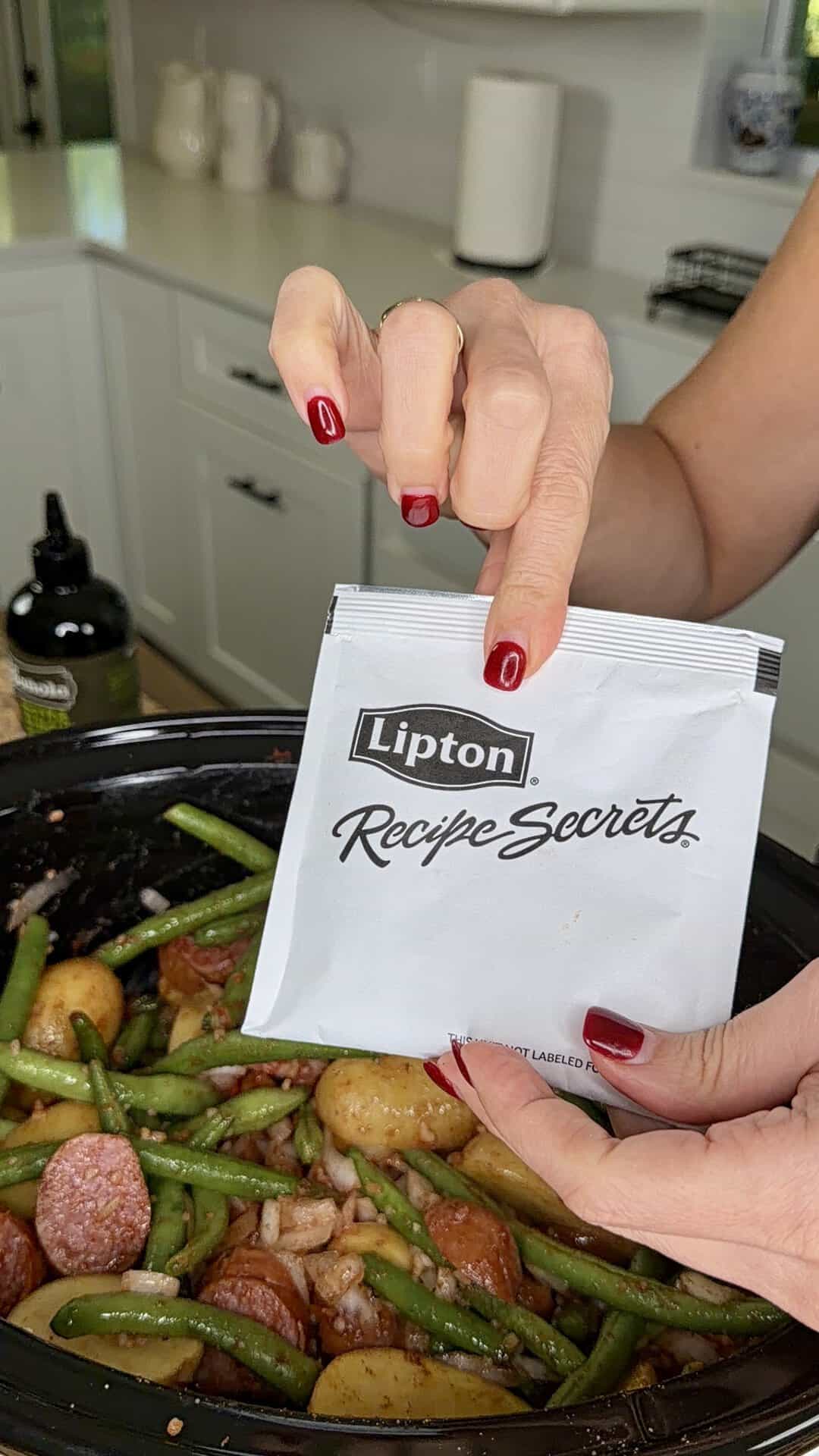 A pair of hands holds a packet of Lipton Recipe Secrets mix over a slow cooker filled with sausage, green beans, and potatoes.