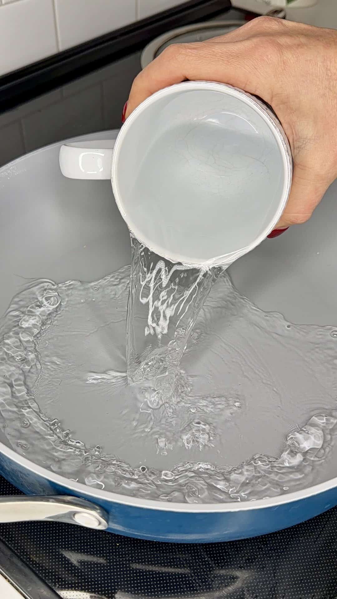 A hand pours water from a white mug into a light-colored frying pan.