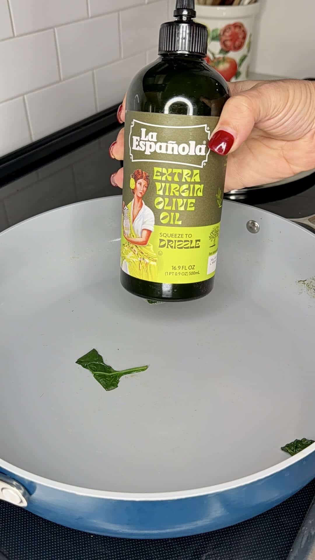 A hand holds a squeeze bottle of La Española extra virgin olive oil over a gray frying pan containing a single leaf of spinach.
