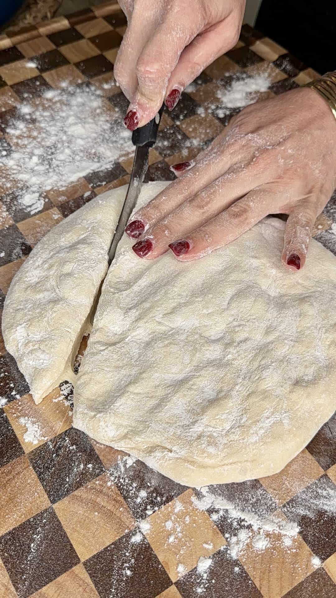 Hands with red nail polish using a knife to cut a floured piece of dough on a checkered wooden board.