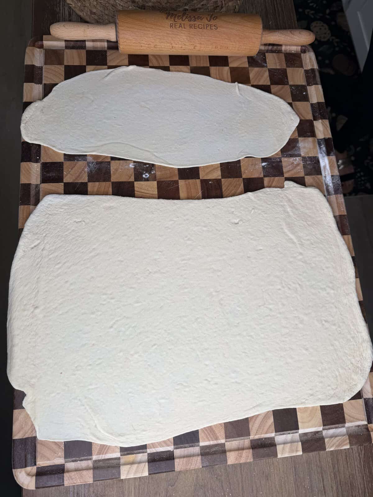 Two pieces of rolled-out dough on a checkered wooden cutting board with a rolling pin at the top.